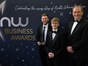 Three people dressed in formal suits standing in front of a dark blue background with writing "NW Business Awards"
