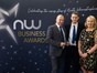 Three people dressed in formal wear, two wearing suits and one wearing a floral print dress, standing in front of a dark blue background with writing "NW Business Awards"