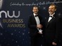Two people dressed in formal suits standing in front of a dark blue background with writing "NW Business Awards"