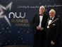 Two people dressed in formal suits standing in front of a dark blue background with writing "NW Business Awards"