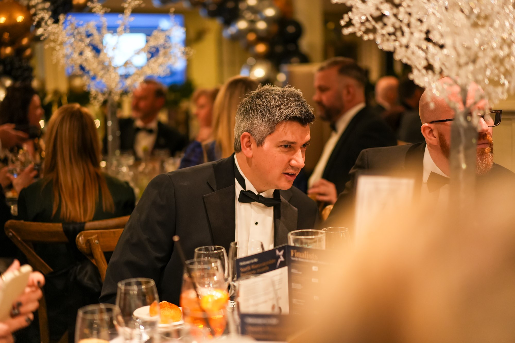 male guest wearing  a black tie evening suit sat at dining table talking with other award evening attendees