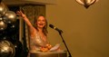 lady stood at a lectern in a silver evening gown, she has a large smile on her face and is pointing out to other people who are attending an awards event evening