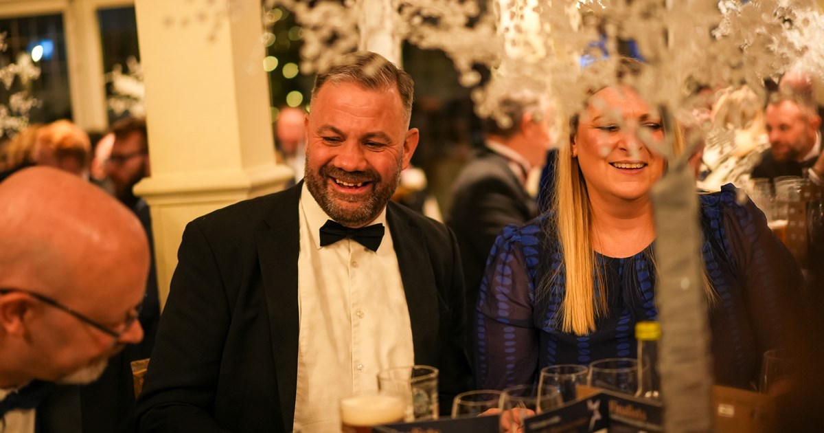 male wearing a black tie evening suit and female wearing an cocktail dress laughing and talking with other award attendees sat at a dining table